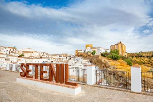 Grandes letras rojas de SETENIL en Setenil de las Bodegas con casas encaladas y viviendas trogloditas en la ladera bajo cielo azul