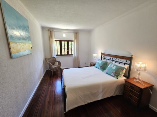 Bright bedroom in Casa Oleias villa, Sesimbra with queen bed, wooden nightstand, wicker chair, and patio door with pool view