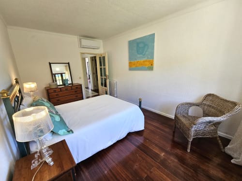 Bright bedroom in Sesimbra villa with white bed, wood dresser, wicker chair, and abstract artwork