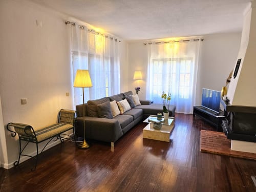 Modern living room in Casa Oleias villa, Sesimbra with grey sofa, wood flooring, TV, and natural light from floor-to-ceiling