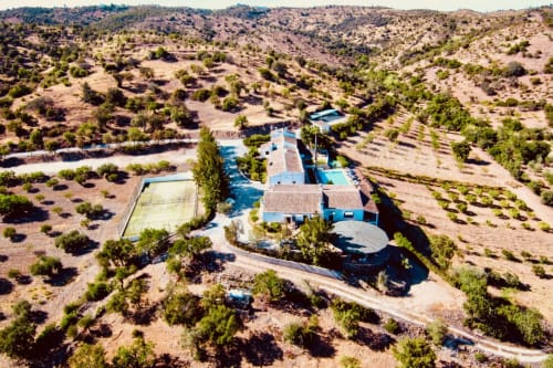 Vista aérea da quinta rural Herdade da Côrte com piscinas azuis, quadra de ténis, cercada por olivais em Tavira,