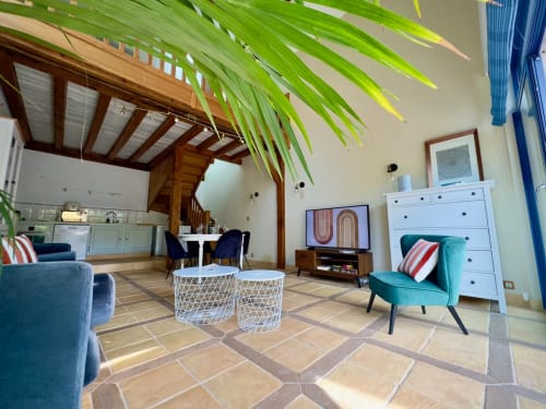 Salon ouvert contemporain à La Maison sous les Vignes, gîte français, avec fauteuil bleu-vert, rangements blancs et poutres en bois