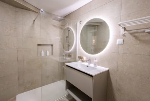 Modern bathroom at J Home Amadora with illuminated oval mirror, white sink vanity, walk-in shower with glass enclosure, and