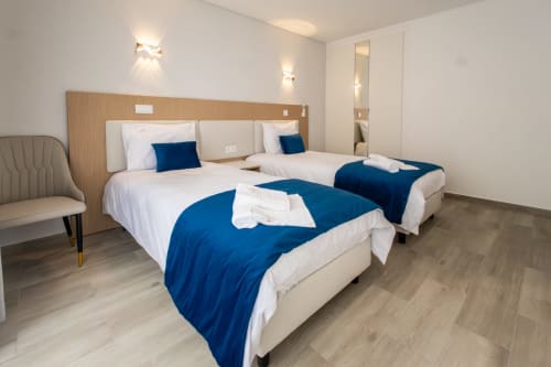 Modern J Home Amadora apartment bedroom with two twin beds, blue throws, wooden headboard, and contemporary lighting.