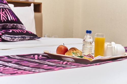 Plateau de bienvenue en chambre à l'Hôtel Yakro avec eau, jus, pomme et pâtisserie sur le lit avec chemin de table à motifs violets