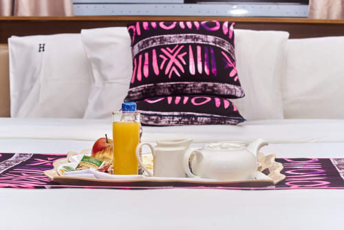 Petit-déjeuner en chambre à l'Hôtel Yakro avec jus, fruits, théière et coussins motifs rose sur le lit