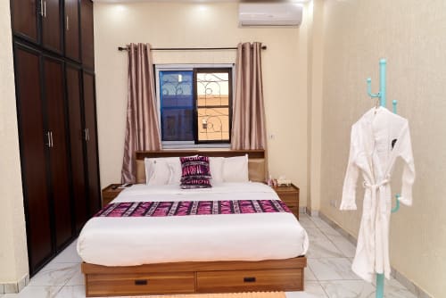 Chambre moderne à l'Hôtel Yakro à Yamoussoukro : lit en bois, draps blancs, chemin de lit violet, climatisation et porte-manteau turquoise.