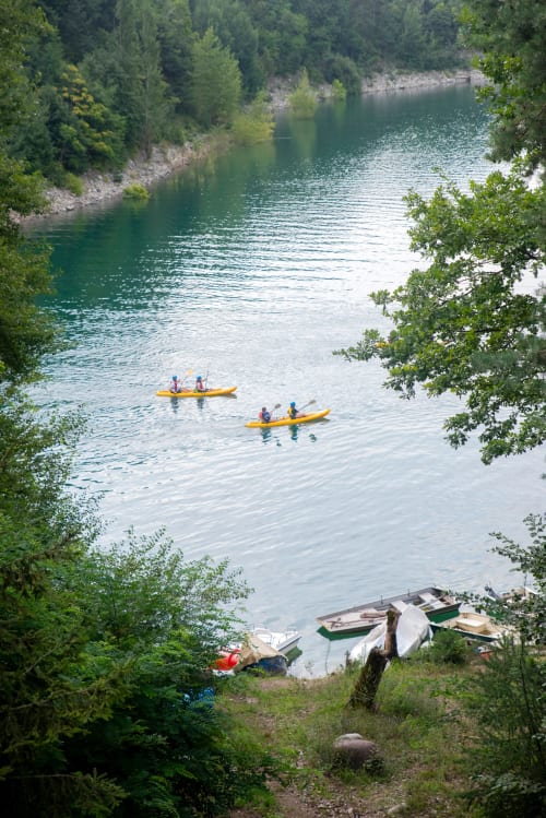 Due kayak remano su un lago turchese vicino a Villa Rosati a Sanzeno, Trentino, con barche ormeggiate e boschi