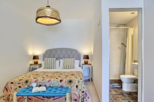 Dormitorio moderno con cabecero tapizado gris, colcha colorida, lámpara colgante y baño privado en Corralejo