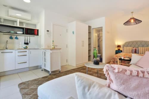 Moderno estudio en Corralejo con cocina abierta, cama con sábanas rosa, muebles blancos y vistas al mar desde el balcón