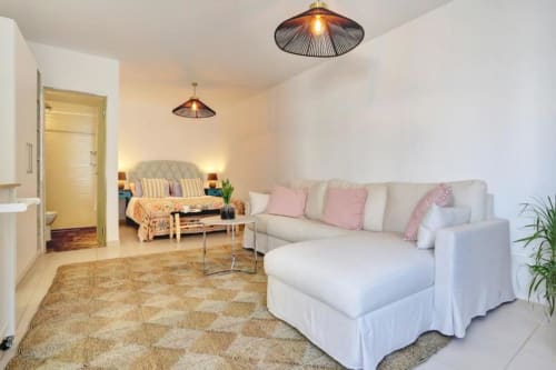 Salón moderno tipo loft en Corralejo con sofá crema, detalles rosa y vistas al mar desde la entrada al dormitorio