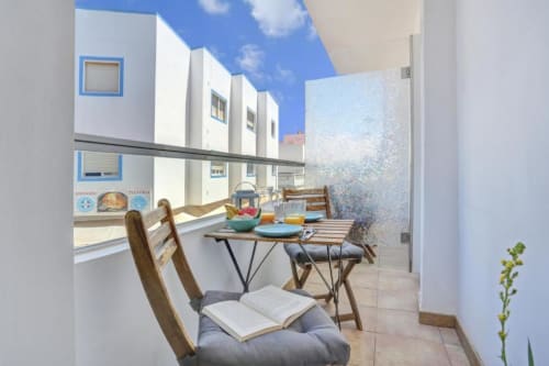 Zona de comedor en balcón del apartamento Kamalahouse SL en Corralejo con vistas al mar, sillas de madera y edificio con detalles azules