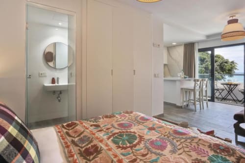 Dormitorio con colcha estampada, baño privado con espejo redondo y vistas al mar desde balcón en apartamento de Corralejo