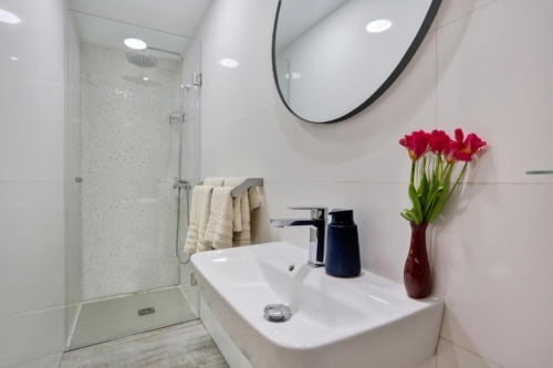 Baño moderno en loft Kamalahouse Corralejo con lavabo blanco, espejo negro, ducha de cristal y flores rojas en florero