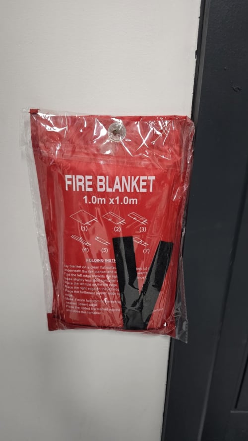 Red fire blanket 1.0m x 1.0m with black handles mounted on white wall at Entrepreneur Stays cottage in Stafford