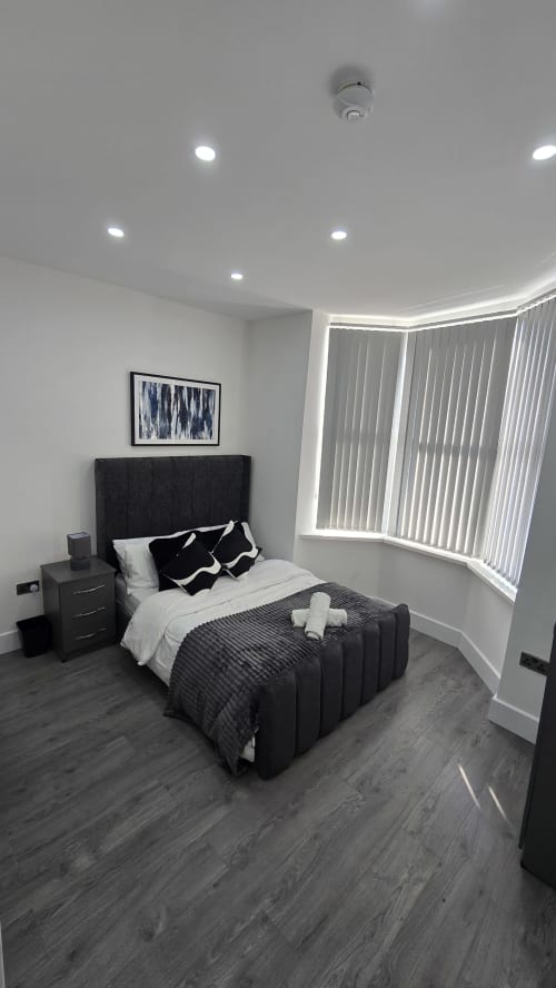 Modern bedroom with black upholstered bed, white bedding, dark wood flooring, and large windows in Stafford cottage rental