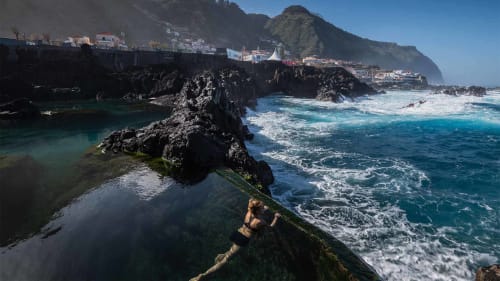 Cove rochosa em Porto Moniz com piscina natural, ondas e edifícios à beira do penhasco sob céu azul