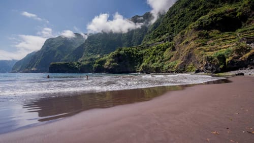 Praia isolada com areia avermelhada, falésias verdejantes e águas tranquilas perto do Refúgio do Nevoeiro em Porto Moniz