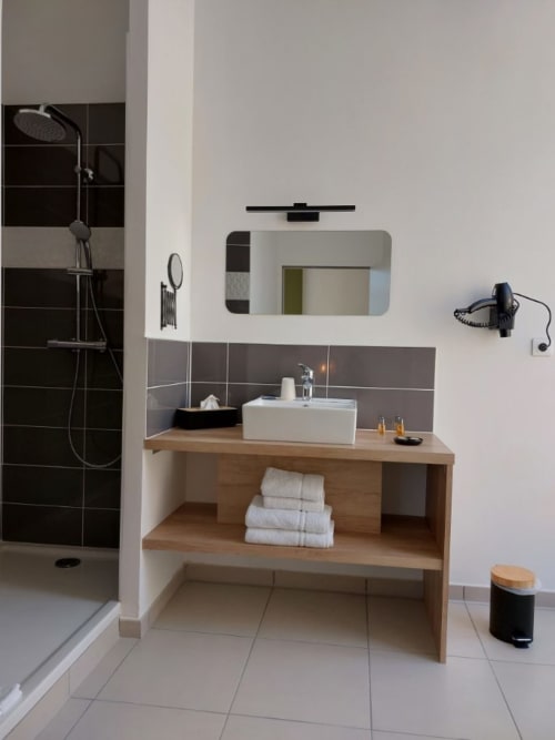 Salle de bain moderne de l'Hôtel Le Château avec meuble vasque en bois, lavabo blanc, douche carrelage noir et miroir éclairant
