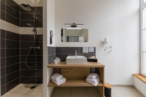 Salle de bains moderne à l'Hôtel Le Château avec douche en carrelage sombre, meuble vasque en bois, lavabo blanc et miroir donnant sur le Rhône