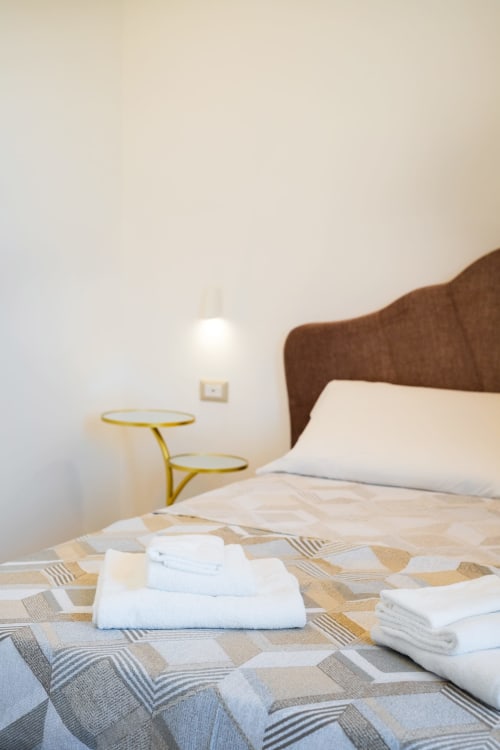 Asciugamani bianchi piegati su copriletto decorato nella camera Eco di Origenes Rooms, Procida, con testiera marrone e tavolino dorato