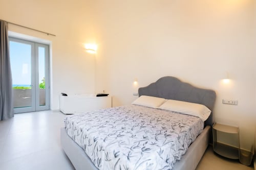 Camera moderna presso Origenes Rooms a Procida con vista mare, testata grigia, biancheria a motivi e accesso al balcone