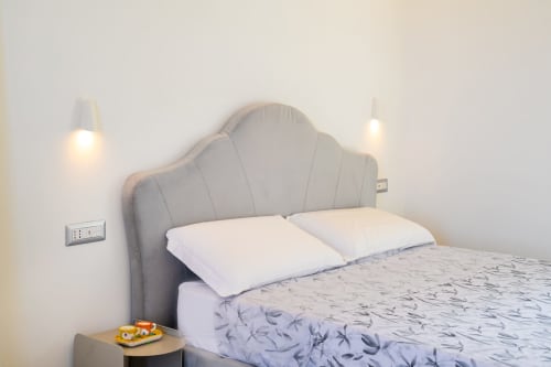 Camera moderna alle Origenes Rooms di Procida con testata imbottita grigia, cuscini bianchi e biancheria decorativa