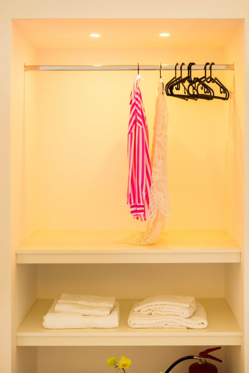 Luminosa cabina armadio presso Origenes Rooms Procida con vestito a righe rosa, scialle in pizzo crema, appendiabiti neri e biancheria piegata sulla mensola