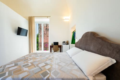 Camera moderna a Procida con balcone con vista sul mare, letto con testata grigia, TV, scrivania e luminosità naturale abbondante