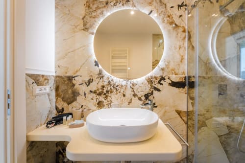 Bagno moderno presso Origenes Rooms, Procida con specchio illuminato rotondo, lavabo bianco e pareti in marmo
