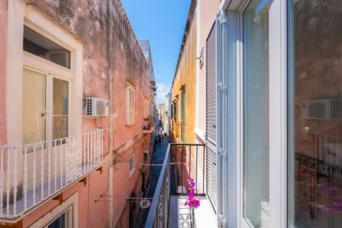 Stretto vicolo colorato a Procida con edifici pastello, balconate bianche e fiori rosa presso Origenes Rooms