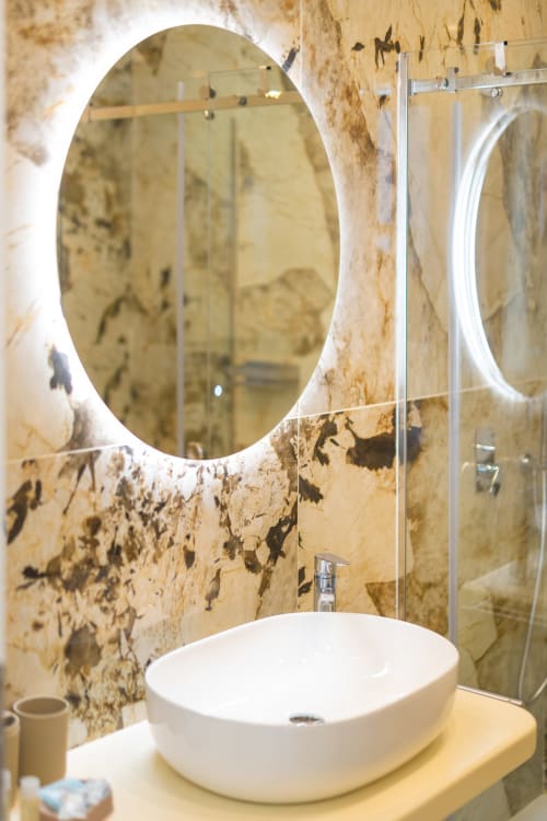 Moderno bagno dell'appartamento Aura presso Origenes Rooms a Procida, con specchio ovale illuminato, lavabo bianco a bacinella e rivestimenti in travertino