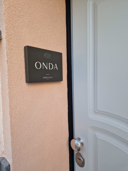 Cartello nero ONDA su muro in stucco color pesca presso Origenes Rooms, Procida, con porta bianca e maniglia in metallo visibili