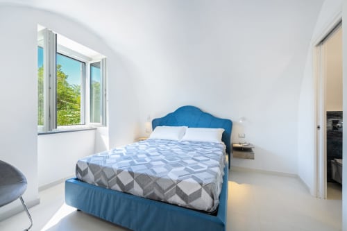Camera da letto luminosa con letto imbottito blu, pareti bianche e finestra con vista sul giardino nell'appartamento di Procida