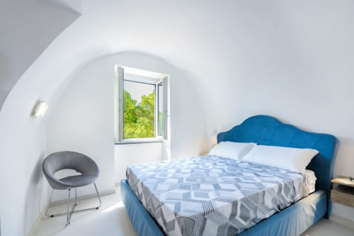 Camera luminosa presso Origenes Rooms, Procida con letto testata blu, pareti bianche arcuate, finestra aperta con vista sulla vegetazione, e