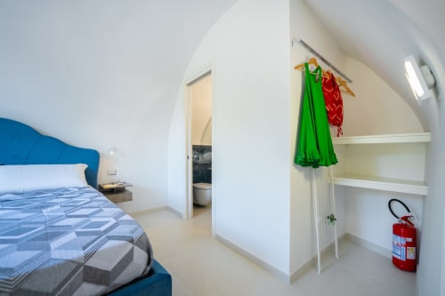 Camera moderna presso Origenes Rooms a Procida con testiera blu, pareti bianche, scaffali aperti, abiti appesi e camino