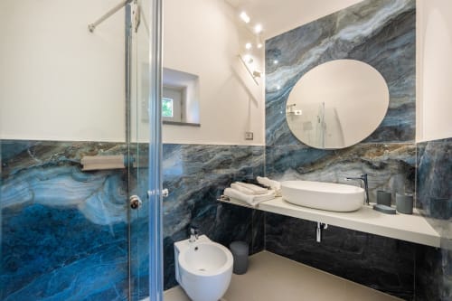 Bagno moderno dell'appartamento Onda presso Origenes Rooms, Procida, con piastrelle in marmo blu, specchio rotondo, lavabo a bacinella e vetro