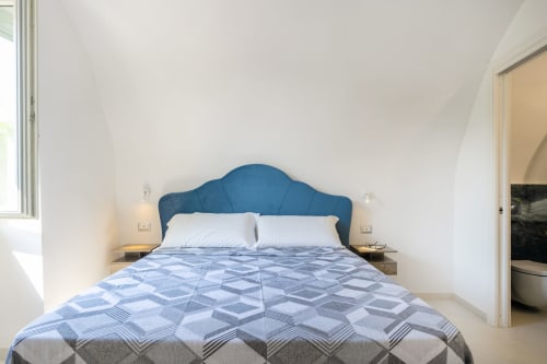 Camera luminosa con testiera trapuntata blu, cuscini bianchi e biancheria con motivi geometrici presso Origenes Rooms a Procida