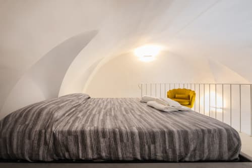Camera moderna negli Origenes Rooms di Procida con soffitto bianco curvo, letto a righe grigie e poltrona gialla con veduta di paesaggio