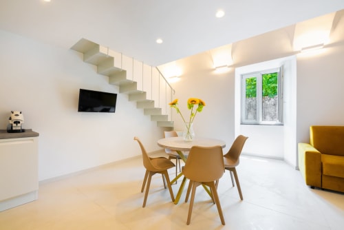 Luminoso appartamento moderno alle Origenes Rooms Procida con zona pranzo, sedie gialle, scale sospese, TV e divano giallo