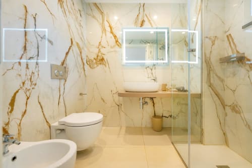 Moderno bagno in marmo a Origenes Rooms, Procida con doppio lavabo, doccia in vetro, WC, specchio riscaldato e eleganti finiture dorate