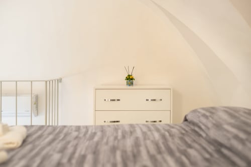 Luminosa camera da letto mansardata alle Origenes Rooms di Procida con pareti color crema, cassettiera moderna, biancheria a righe e accesso alla vista dei monumenti