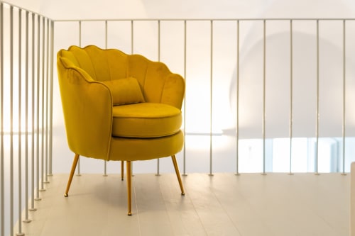 Poltrona curva giallo senape con gambe in metallo nella camera moderna di Origenes Rooms, Procida