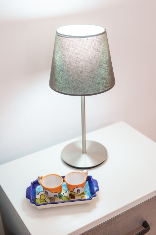 Modern table lamp with gray linen shade and decorative ceramic cups on white nightstand at Origenes Rooms, Procida