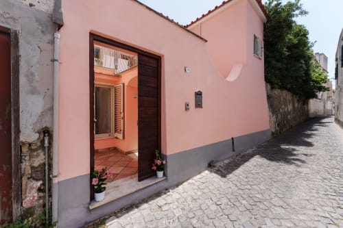Pink and gray exterior of Origenes Rooms B&B in Procida, Italy, with open doorway and flowering plants along the cobblestone