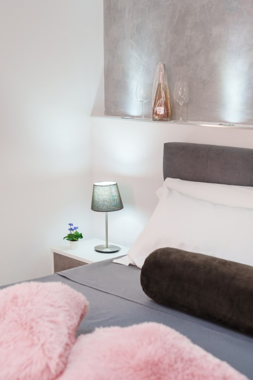 Modern bedroom at Origenes Rooms, Procida with gray headboard, white bedding, blue lamp, and wine glasses on shelf