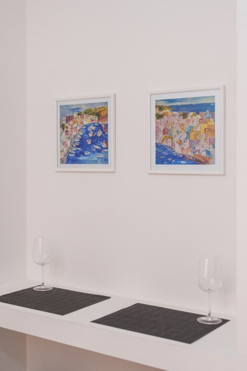 Modern dining setup at Origenes Rooms Procida with framed Mediterranean coastal artwork and wine glasses on white table