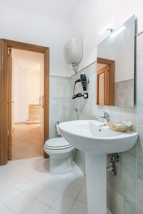 Modern bathroom at Origenes Rooms in Procida with pedestal sink, toilet, mirror, and wooden doors leading to bedroom