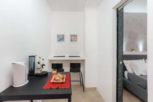 Modern bedroom at Origenes Rooms in Procida with desk, coffee maker, two black chairs, and bed visible through doorway