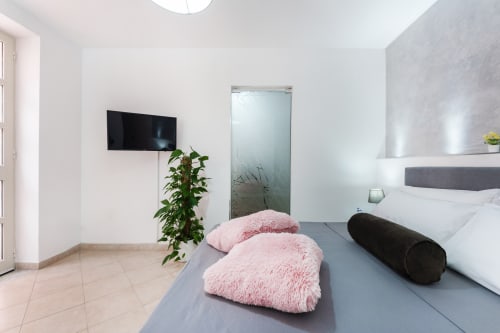 Modern bedroom at Origenes Rooms in Procida with queen bed, pink pillows, wall-mounted TV, and ensuite bathroom with glass